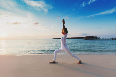 Woman with arms raised exercising at beach against sky