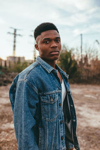 Young serious african american male with modern haircut in denim jacket looking at camera under cloudy sky in fall