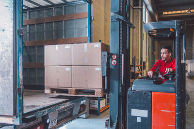 Male driver of forklift truck during shipment packages from big truck in modern warehouse