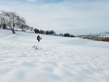 Woman with snowboard walking on snow covered field against sky