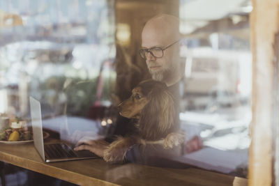 Bald man using laptop while sitting with dog at cafe seen through window