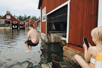 Rear view of father jumping in lake while daughter with smart phone sitting by house