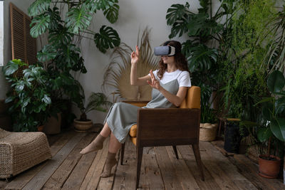 Impressed woman using virtual reality glasses touching air, resting in cozy tropical indoor garden