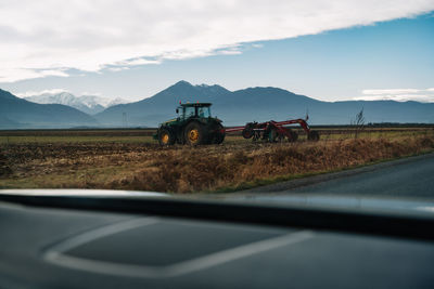 Vehicle on road by agricultural tractor on field against mountain lansdcape and sky during winter