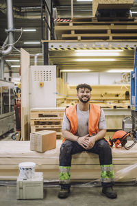 Full length of young carpenter sitting by hardhat in warehouse while looking away