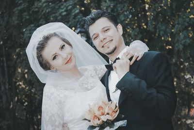Portrait of happy young bride and groom standing against trees