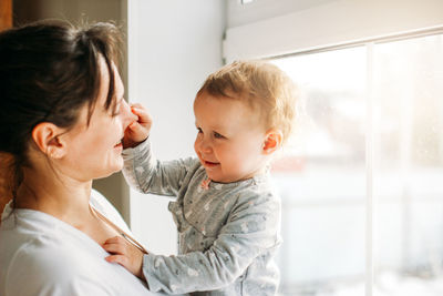 Mother carrying playful daughter while standing against window