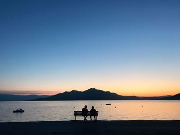 Silhouette people sitting on bench by lake against sky during sunset