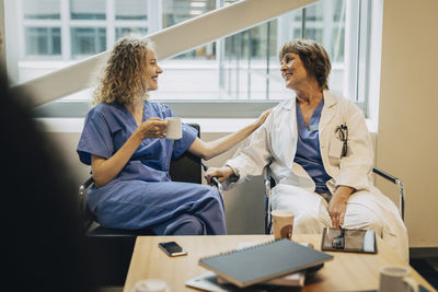 Smiling young nurse talking to mature physician while sitting on chair during coffee break