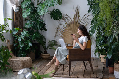 Joyful woman sitting at workplace with laptop and recording voice message surrounded by plants.
