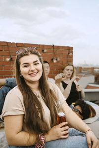 Portrait of smiling young woman holding beer bottle sitting in front of friends on rooftop