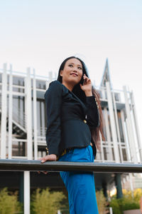 Low angle view of young woman standing on railing against sky