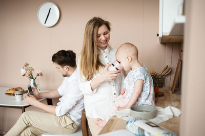 Mother feeds child sitting on kitchen table while father chats online.