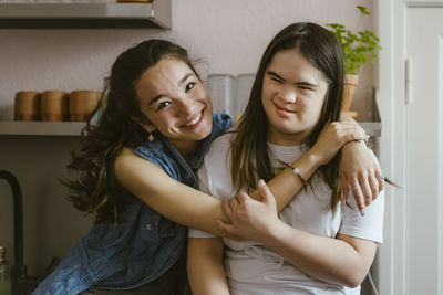 Portrait of smiling young woman hugging sister with down syndrome in kitchen at home