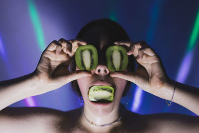 Close-up of woman covering eyes with kiwi slices against lights on wall