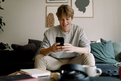 Happy teenage boy using mobile phone while sitting on sofa at home