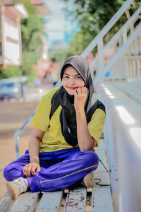 Portrait of smiling young woman wearing hijab sitting on bench in city