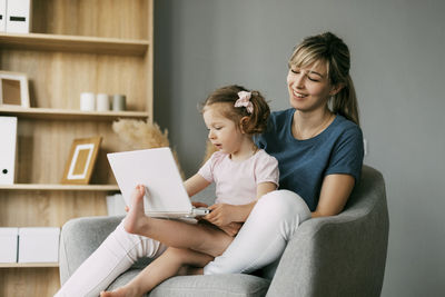 A little girl and her mom watch cartoons on a laptop while sitting in a chair together