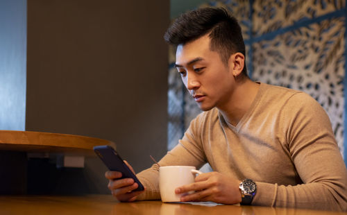 Thoughtful asian male relaxing in armchair in cozy coffee shop and enjoying hot beverage while browsing smartphone