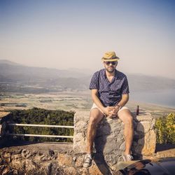 Full length of man sitting on stone railing over mountain against sky