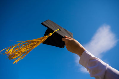 Cropped hand holding mortarboard against clear blue sky