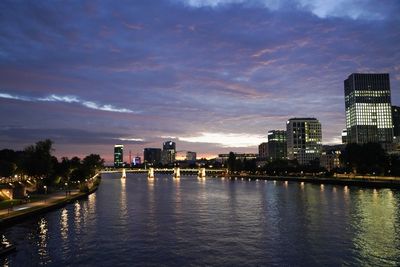 River by illuminated buildings against sky at sunset