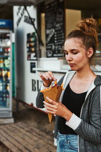 Teenage girl eating potato fries from carton cone standing in front of food truck