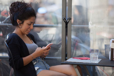 Young woman using mobile phone while sitting by window at restaurant