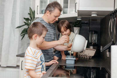 Dad helps to prepare pie or muffin or pancake dough for two sons in the kitchen at home. 