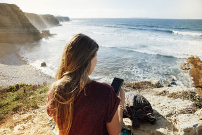 Rear view of woman using mobile phone while sitting on cliff by sea