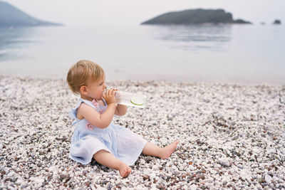 Full length of girl drinking lemonade sitting on pebbles at beach