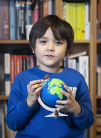 Portrait of boy holding globe against bookshelf