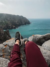 Low section of person relaxing on rocks by sea against sky