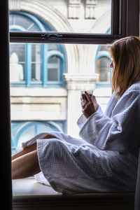 Side view of woman having coffee while sitting on window sill