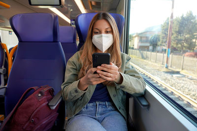 Student girl wearing protective medical mask on train enjoying using smartphone app