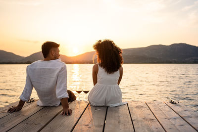 Rear view of couple sitting on shore against sea during sunset