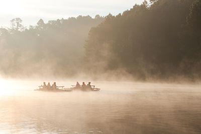 Silhouette people on boats in river against trees during foggy sunrise