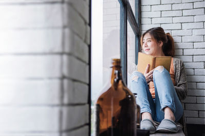 Smiling young woman with book sitting by window at home