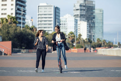 Friend walking by young woman riding push scooter in city