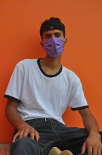 A indian young guy wearing face mask, looking at camera sitting against orange wall with copy space 