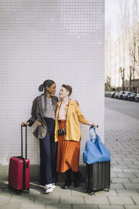 Full length of smiling lesbian couple with wheeled luggage standing against wall