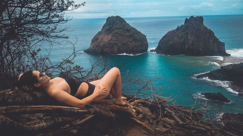 Sensuous woman lying on rock by sea against sky