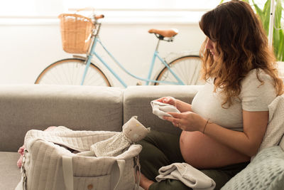 Pregnant mid adult woman looking baby clothing while sitting on sofa at home