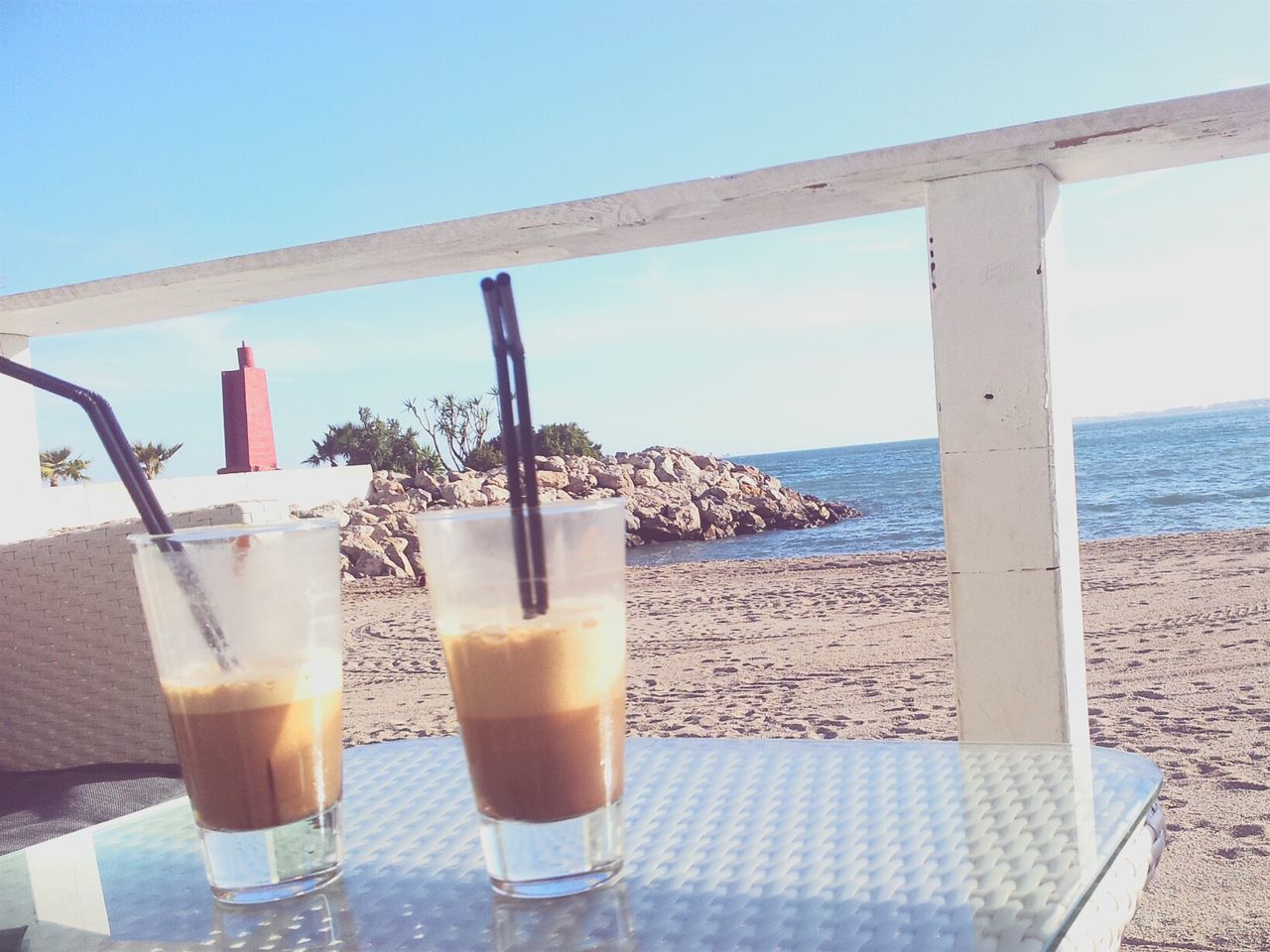 drink, sea, food and drink, horizon over water, refreshment, water, table, beach, freshness, drinking glass, sky, sunlight, drinking straw, close-up, clear sky, still life, blue, shore, outdoors, day