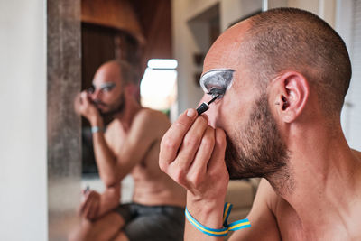 Adult shirtless bearded male transgender with eyebrow makeup and cosmetic lens applying black eyeliner