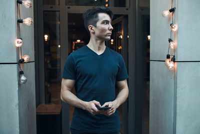 Young man standing in front of restaurant in the city center holding smartphone looking away