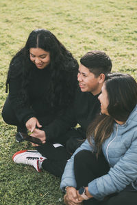 Hispanic teenager holding smartphone looking away while sitting on grass with mother and sister 