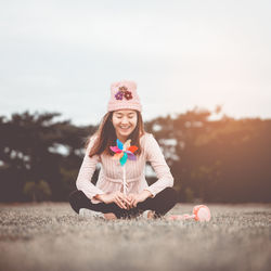 Smiling woman with closed eyes holding pinwheel while sitting on grassy field against clear sky