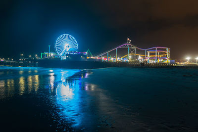 Illuminated ferris wheel by sea against sky at night