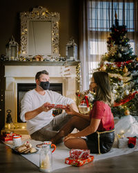 Woman wearing mask giving gift to man by christmas tree